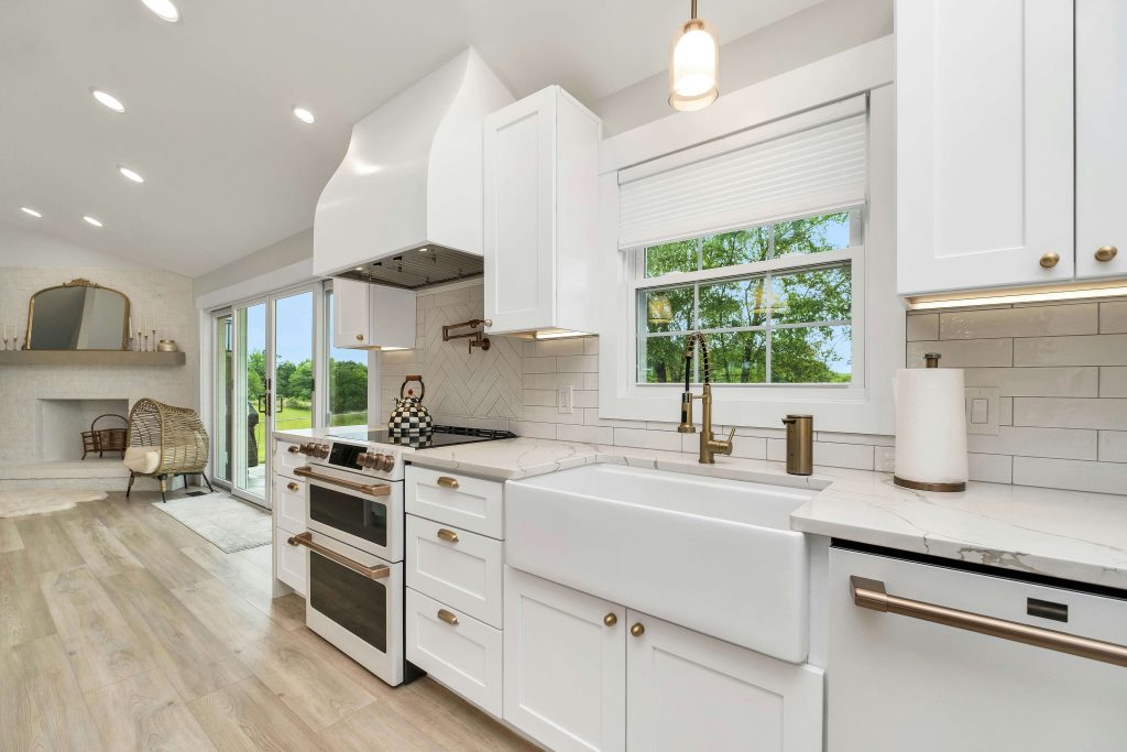 Remodeled kitchen with white shaker cabinets, quartz countertops, farmhouse sink, light hardwood floors, and gold hardware
