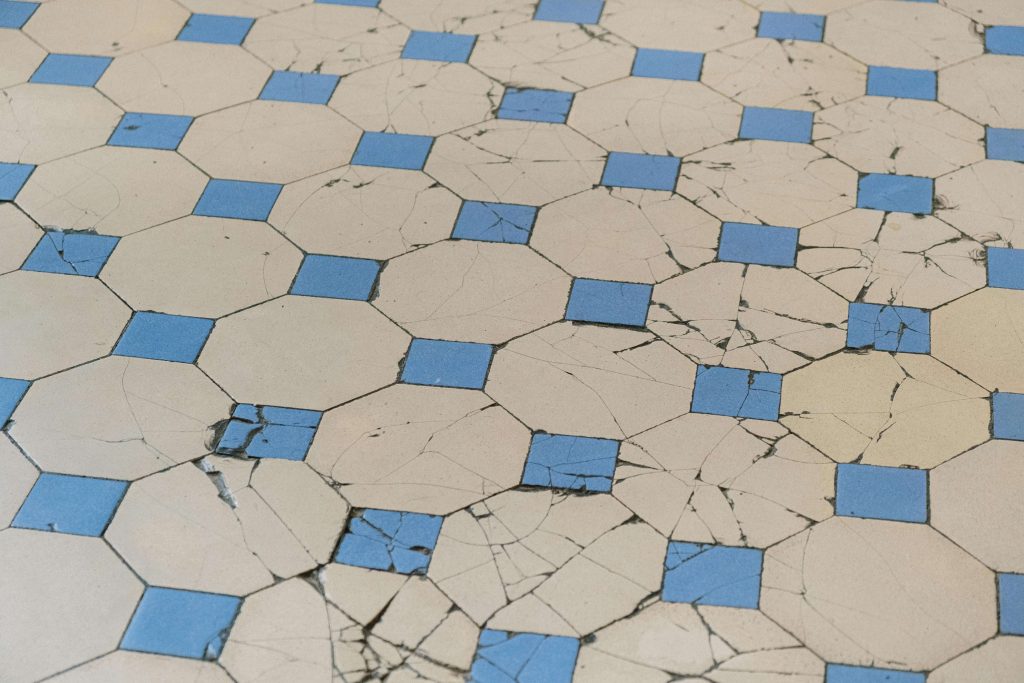 Geometric tile floor pattern showing visible cracks in beige tiles with blue accent tiles, demonstrating tile cracking from slab movement and reflective cracking