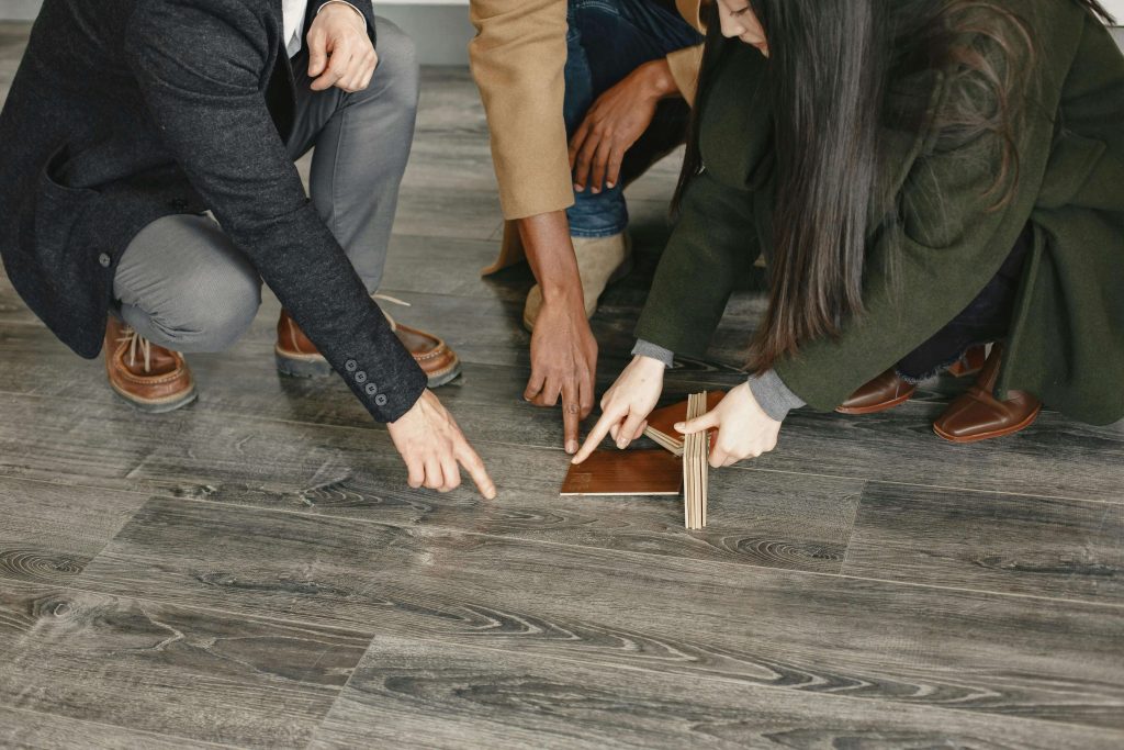 Three people crouching on a gray hardwood floor examining a wood flooring sample for installation issues